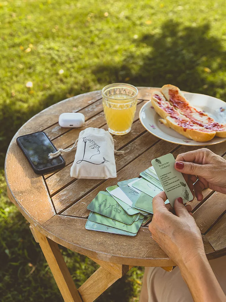 Femme triant des cartes vertes sur une table de jardin, avec écouteurs, jus et tartine - collection Rémission Toujours L pour femme après un cancer