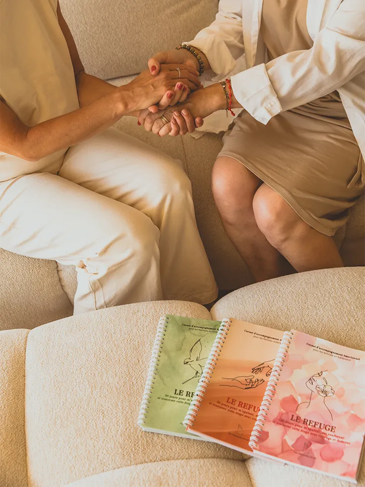 Deux femmes se tenant les mains en signe de soutien, avec les carnets Le Refuge des trois collections posés à côté - accompagnement Toujours L pour femme atteinte de cancer et femme aidante