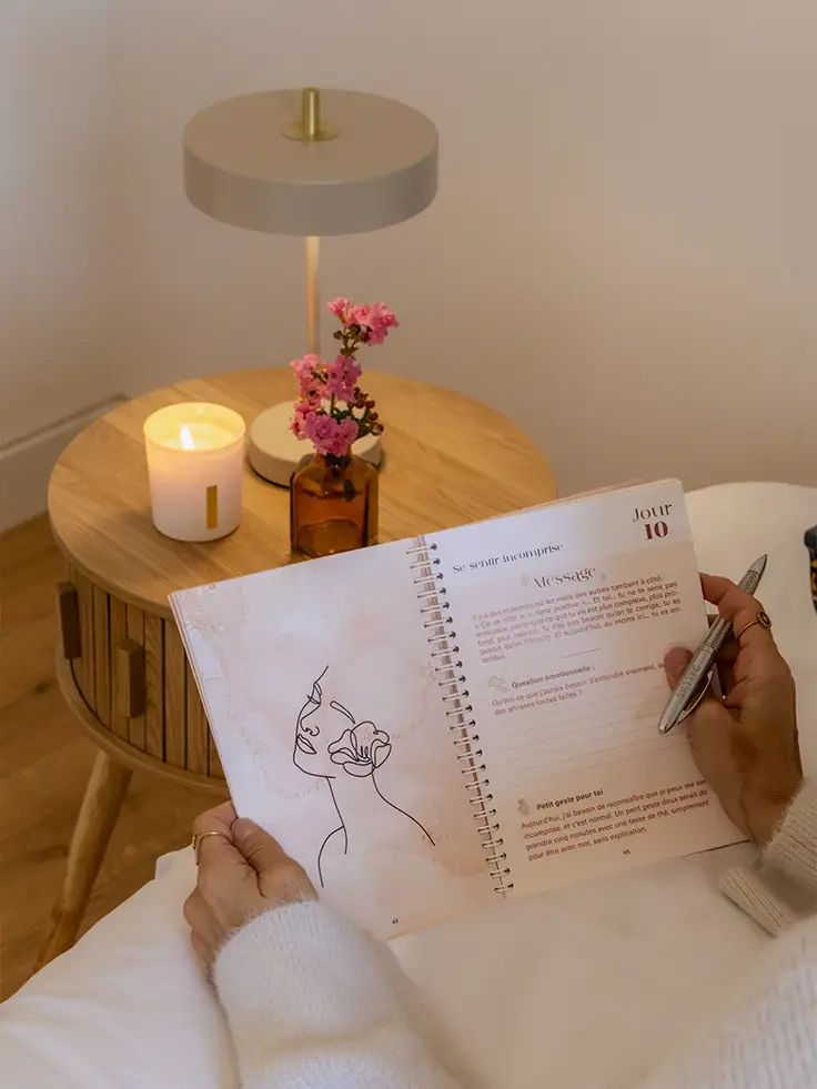 Femme écrivant dans un carnet ouvert près d'une bougie et de fleurs sur un guéridon - collection Traitement Toujours L pour femme atteinte de cancer