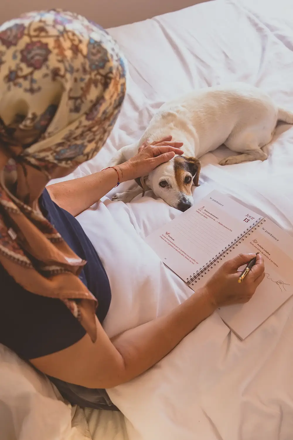 Femme portant un foulard caressant son chien allongé sur un lit, avec un carnet ouvert à côté - collection Traitement en vente sur Toujours L, site pour femme atteinte de cancer