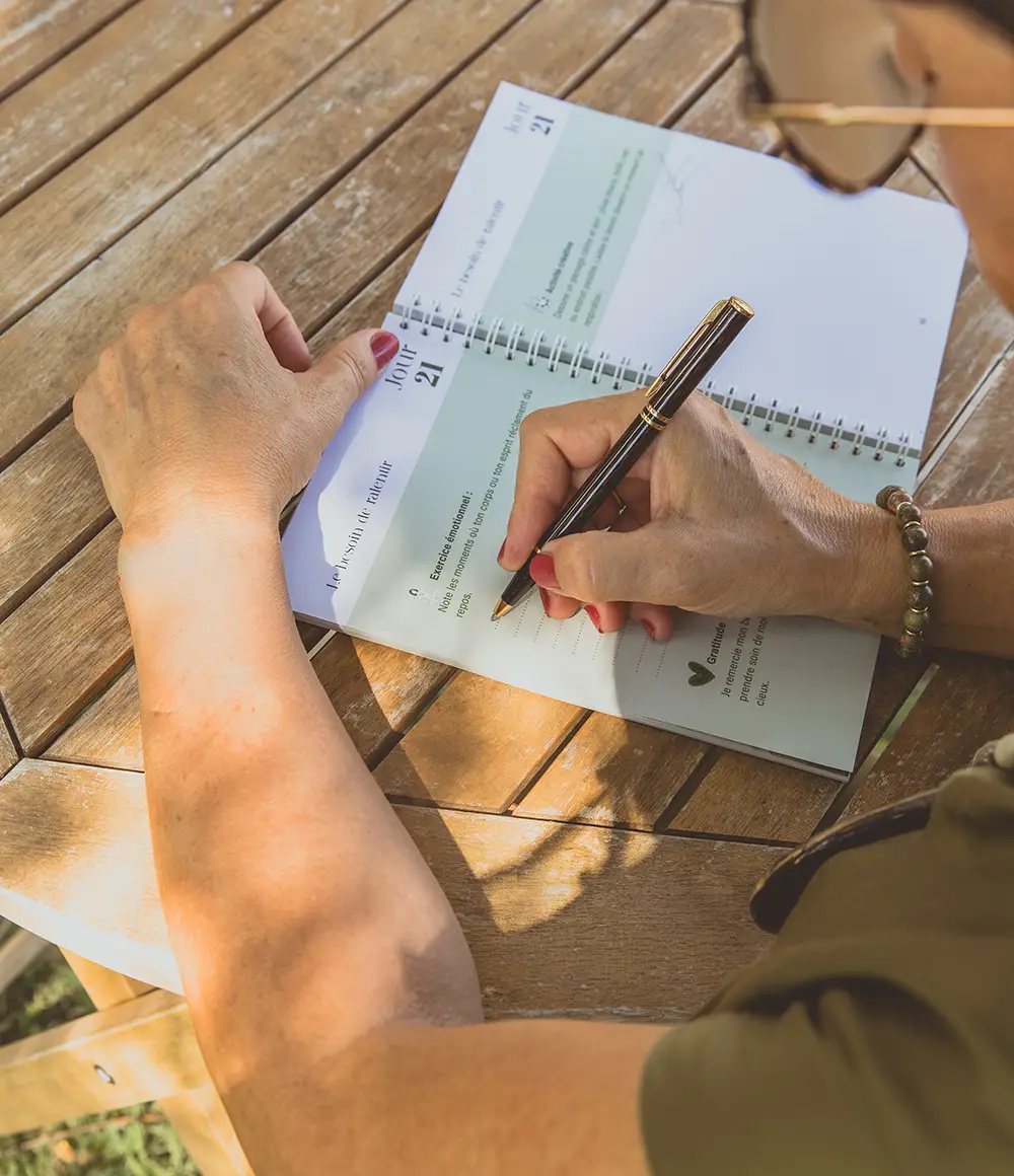 Femme écrivant dans un carnet en extérieur sur une table en bois - collection Rémission en vente sur Toujours L, site pour femme atteinte de cancer