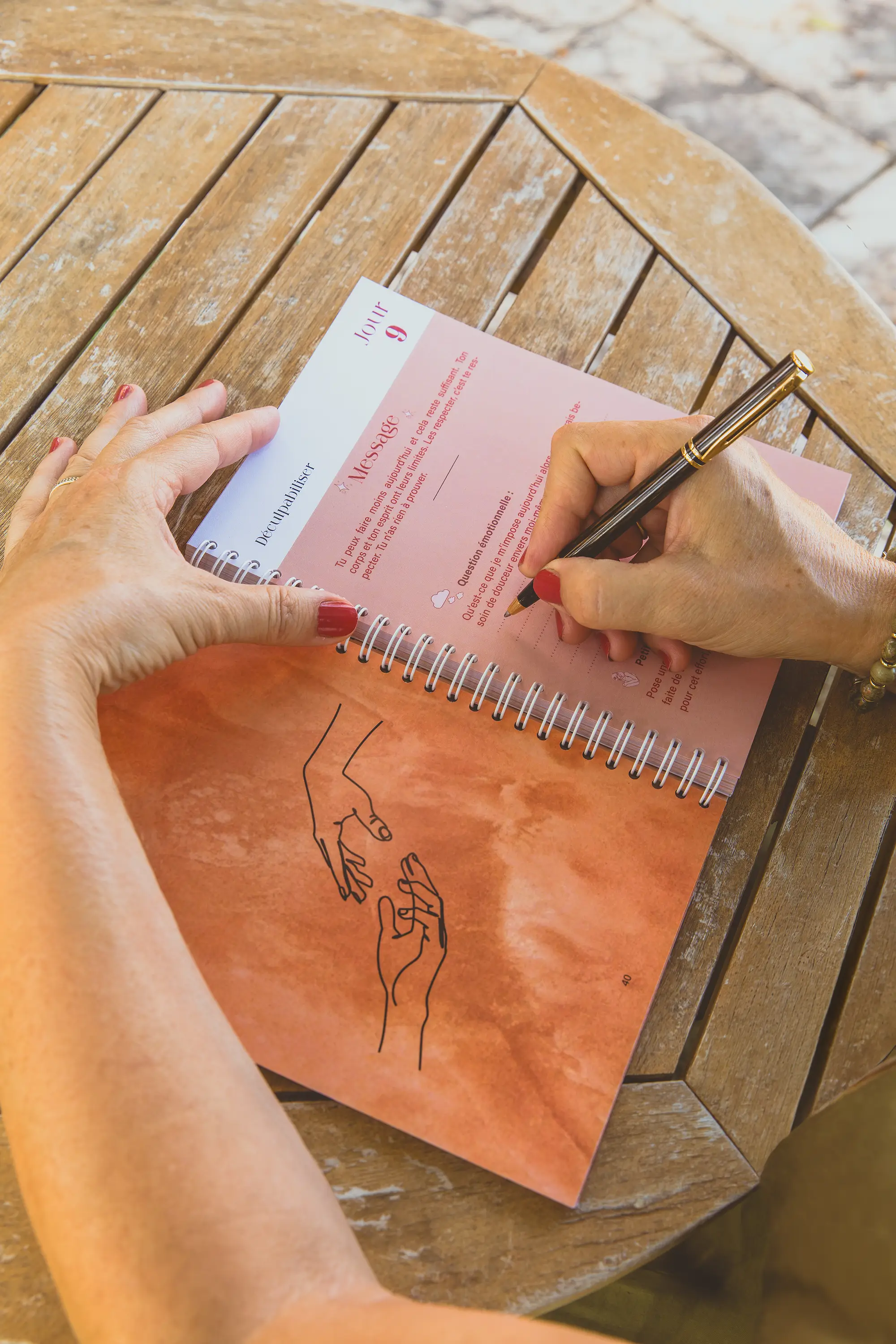 Femme écrivant dans un carnet aux pages roses sur une table en bois extérieure - collection Traitement en vente sur Toujours L, site pour femme atteinte de cancer
