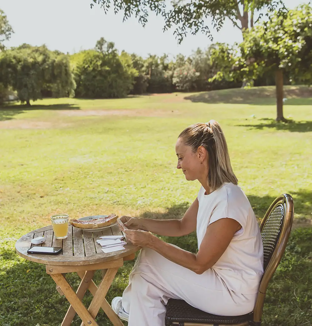 Femme assise seule à une table de jardin en extérieur, avec un carnet vert ouvert, une tartine et un jus - collection Rémission en vente sur Toujours L, site pour femme après un cancer