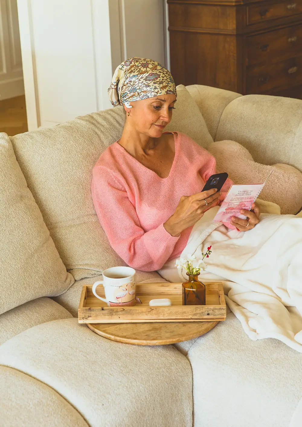 Femme portant un foulard, assise sur un canapé, lisant un carnet aux pages roses avec un plateau petit-déjeuner à côté - collection Traitement en vente sur Toujours L, site pour femme atteinte de cancer