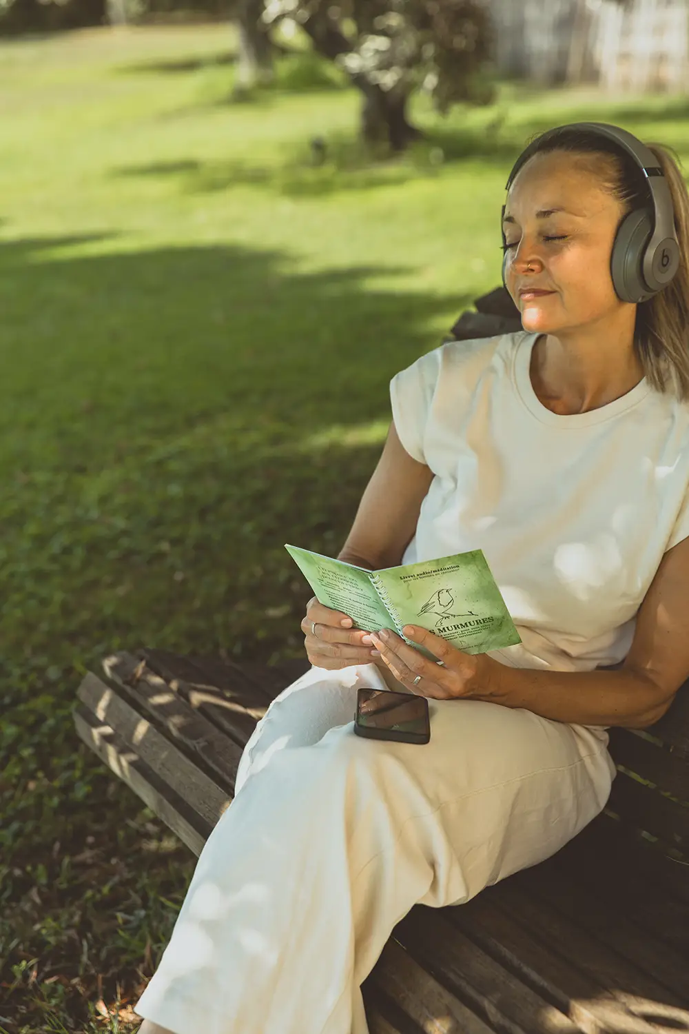 Femme au casque assise sur un banc, lisant un livret vert Les Murmures dans un parc - collection Rémission en vente sur Toujours L, site pour femme après un cancer