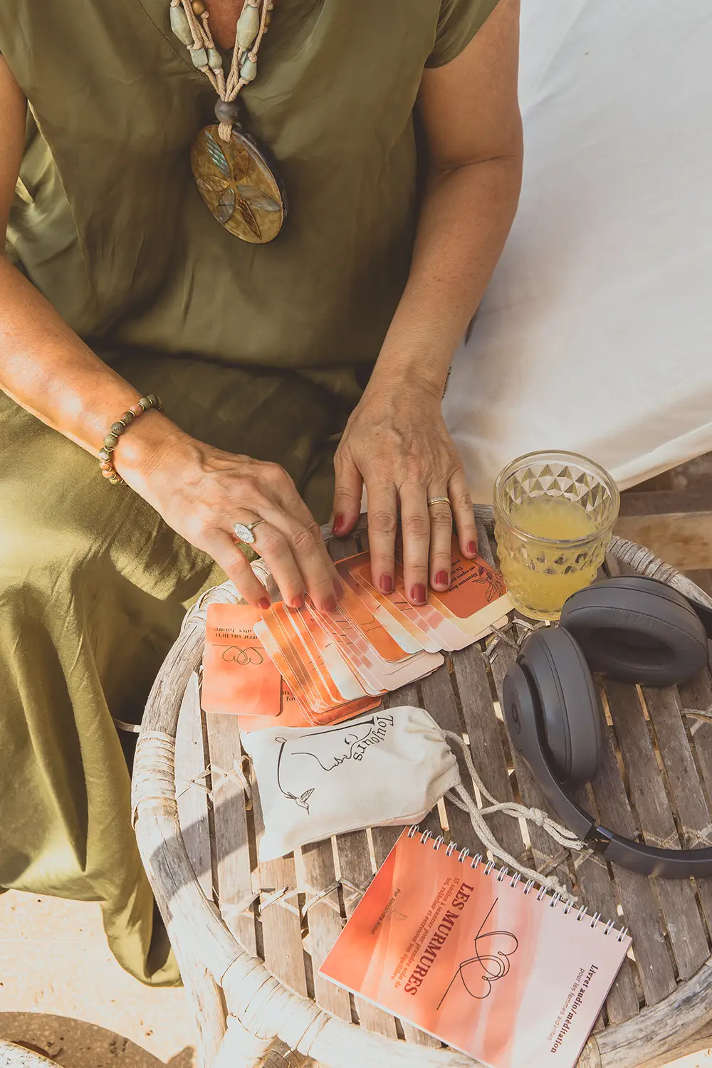 Femme en robe verte triant des cartes orangées sur une table en bois, avec un livret Les Murmures, un casque et un jus - collection Aidantes en vente sur Toujours L, site pour femme aidante d'une proche atteinte de cancer