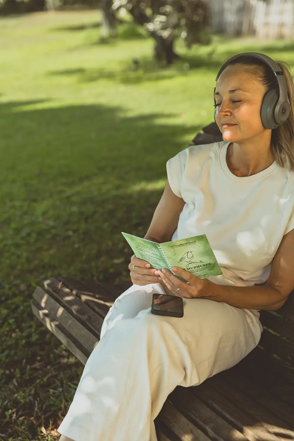 Femme au casque assise sur un banc, lisant un livret vert Les Murmures dans un parc - collection Rémission en vente sur Toujours L, site pour femme après un cancer