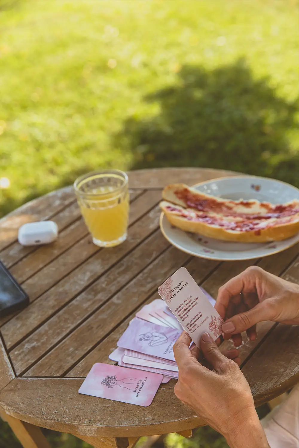 Femme triant des cartes roses sur une table de jardin, avec écouteurs, jus et tartine - collection Traitement en vente sur Toujours L, site pour femme atteinte de cancer