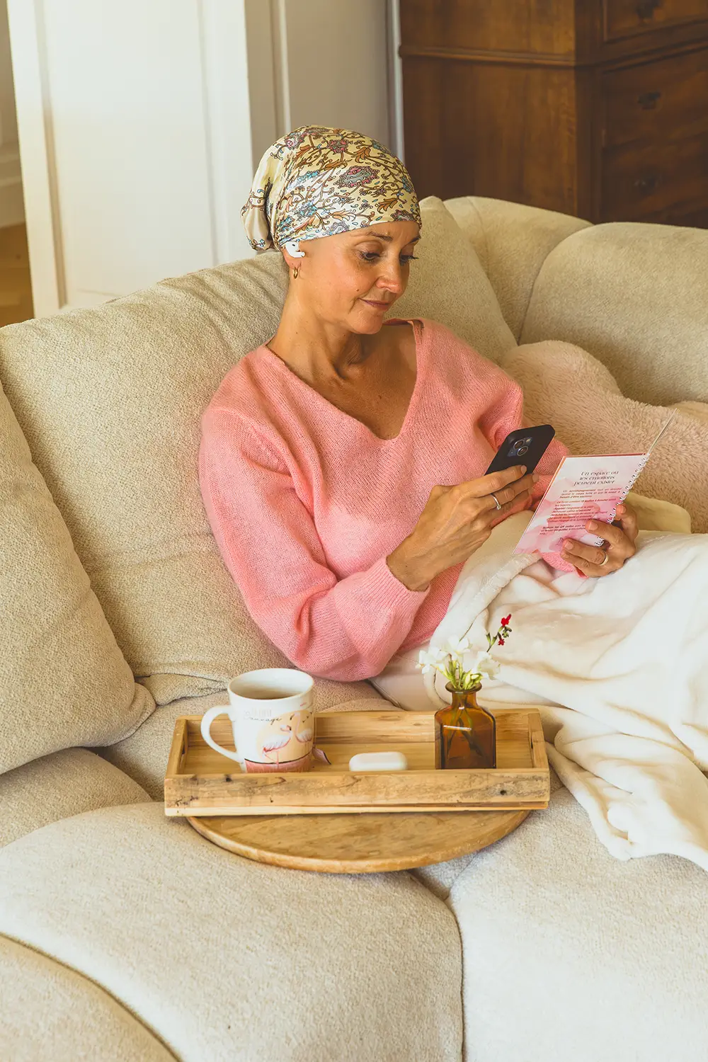 Femme portant un foulard, assise sur un canapé, lisant un carnet rose avec un plateau petit-déjeuner à côté - collection Traitement en vente sur Toujours L, site pour femme atteinte de cancer